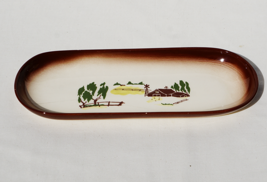 Cream colored oblong ceramic dish with brown border. It is decorated with a painted country scene that includes a barn, windmill, and trees. 